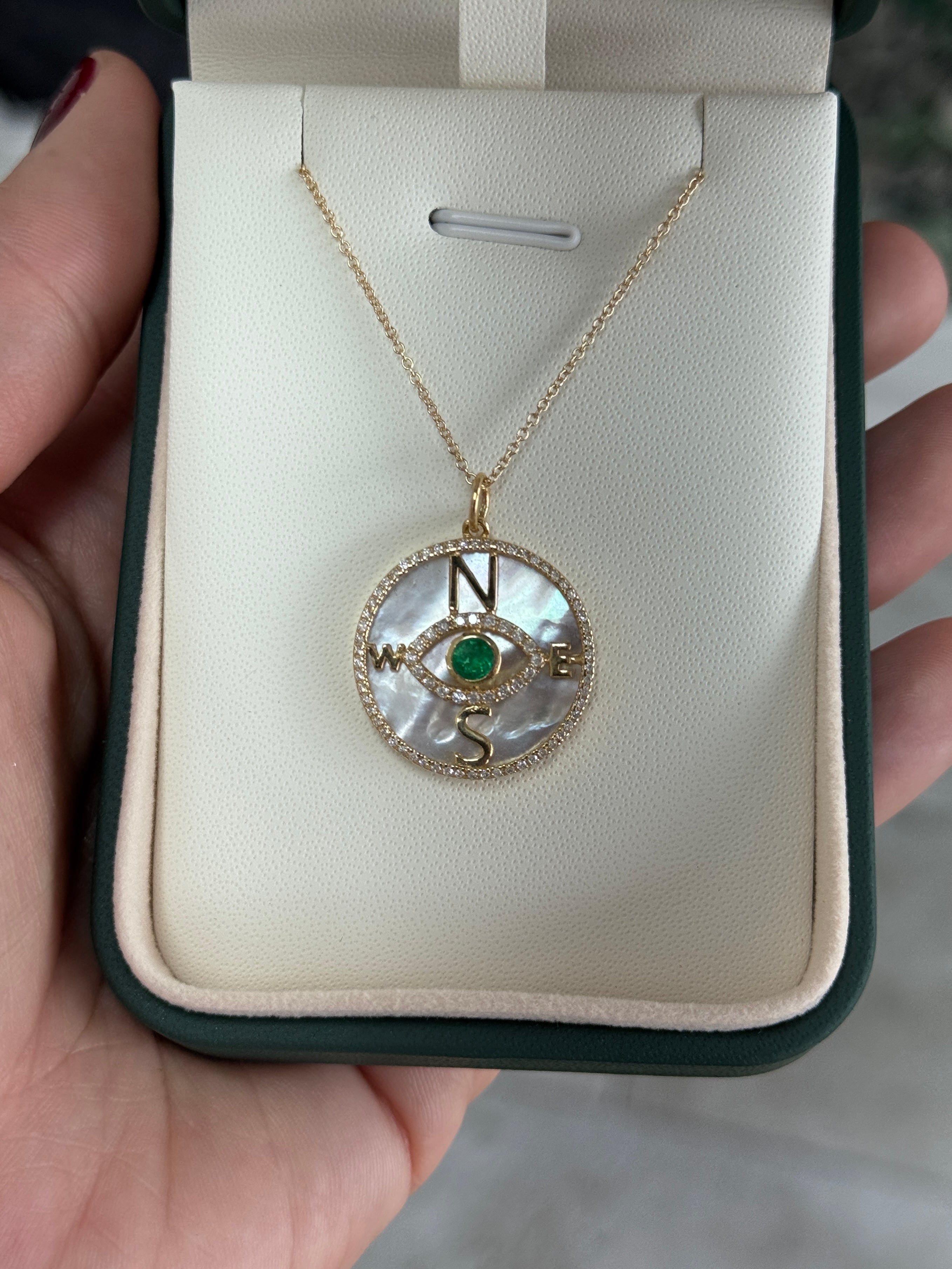14K gold emerald and diamond compass evil eye pendant necklace with mother of pearl background, featuring natural round emeralds and diamonds.