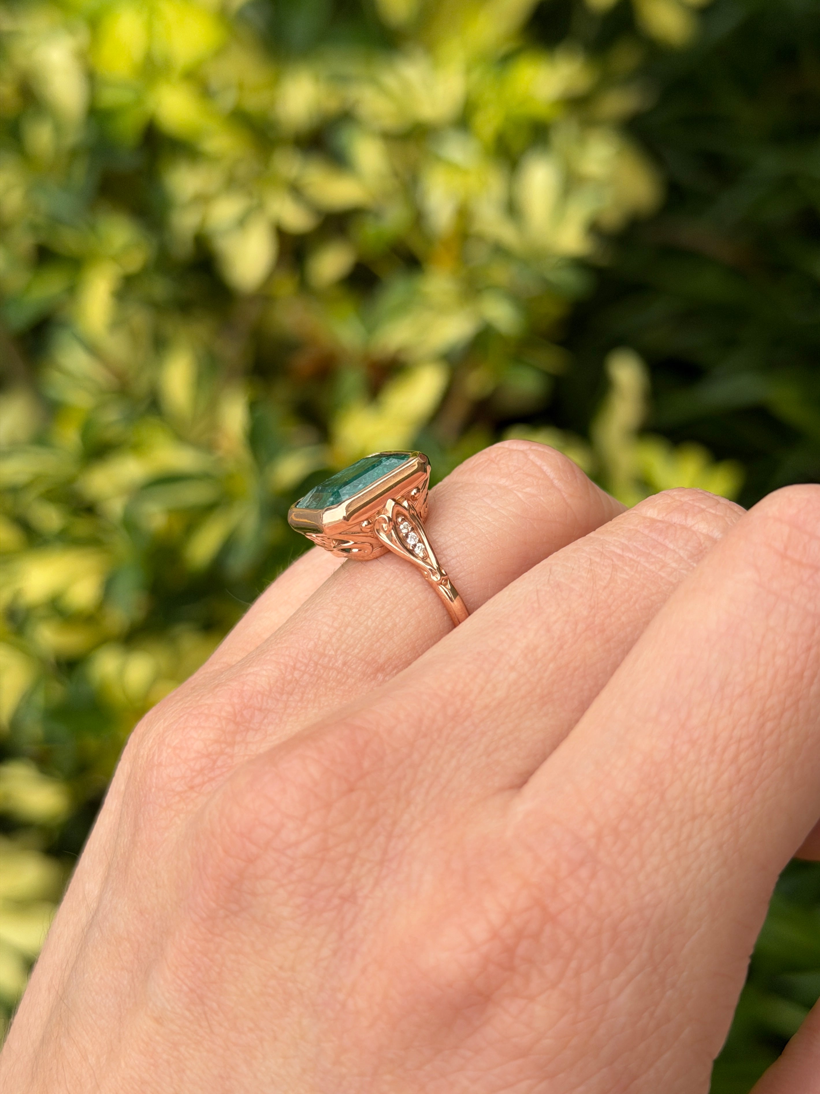 Vintage-inspired 14K rose gold emerald bezel ring with diamond shank and scroll basket detail, featuring 6.20tcw of emeralds and diamonds.