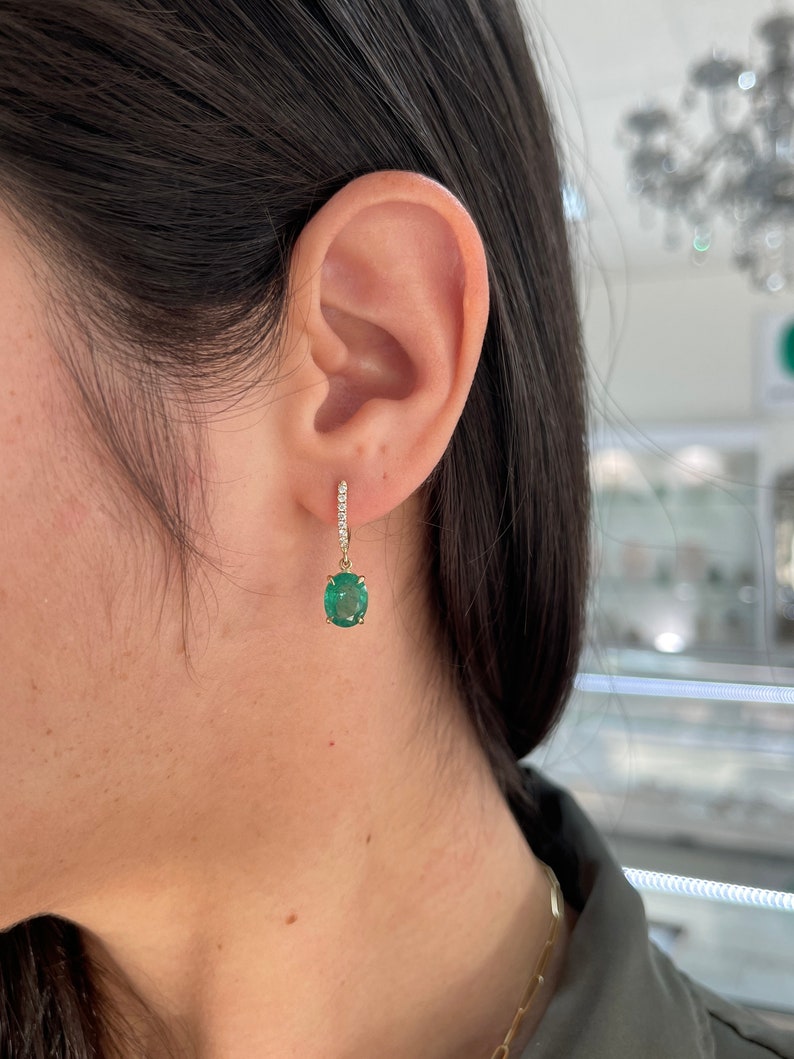 14K gold huggie dangle earrings featuring dark green oval cut emeralds and diamond accents.