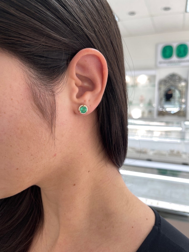 14K gold emerald and diamond halo stud earrings featuring 8.5mm round Colombian emeralds and sparkling diamonds.