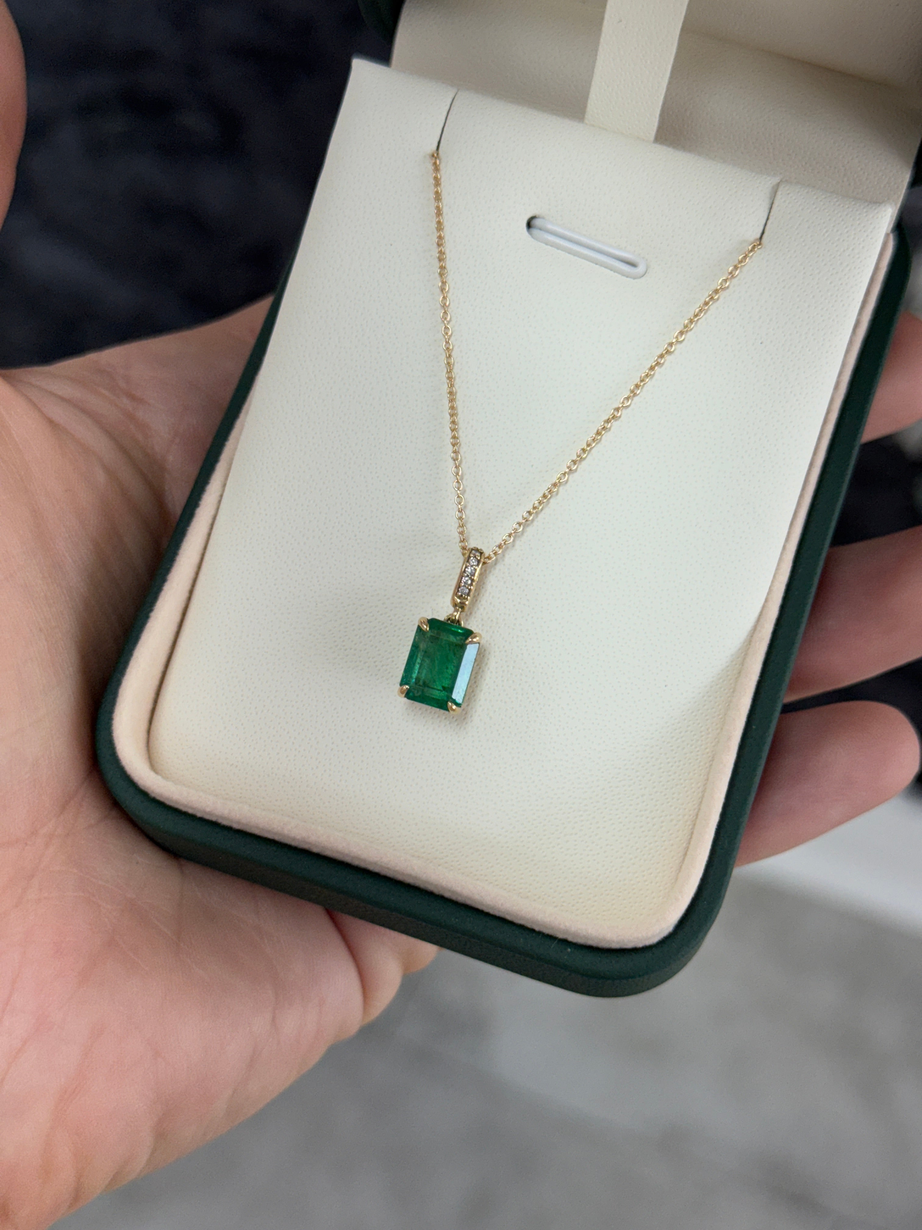 Emerald cut emerald pendant with diamond bail in 14K gold, displayed on a jewelry stand with a black background.