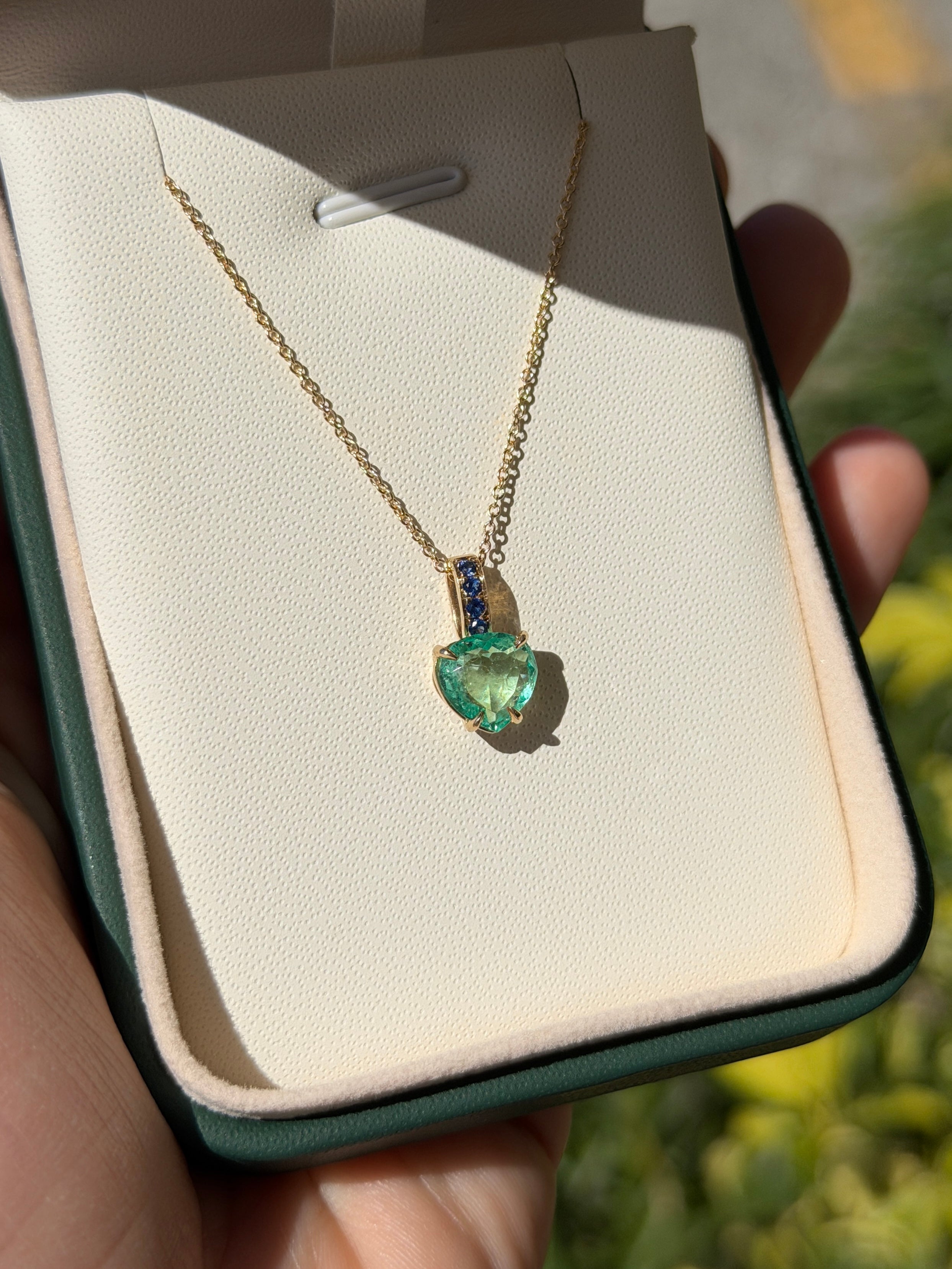 Colombian pear-shaped emerald pendant with blue sapphire bail in 14K gold, featuring a heart-shaped design and intricate craftsmanship.