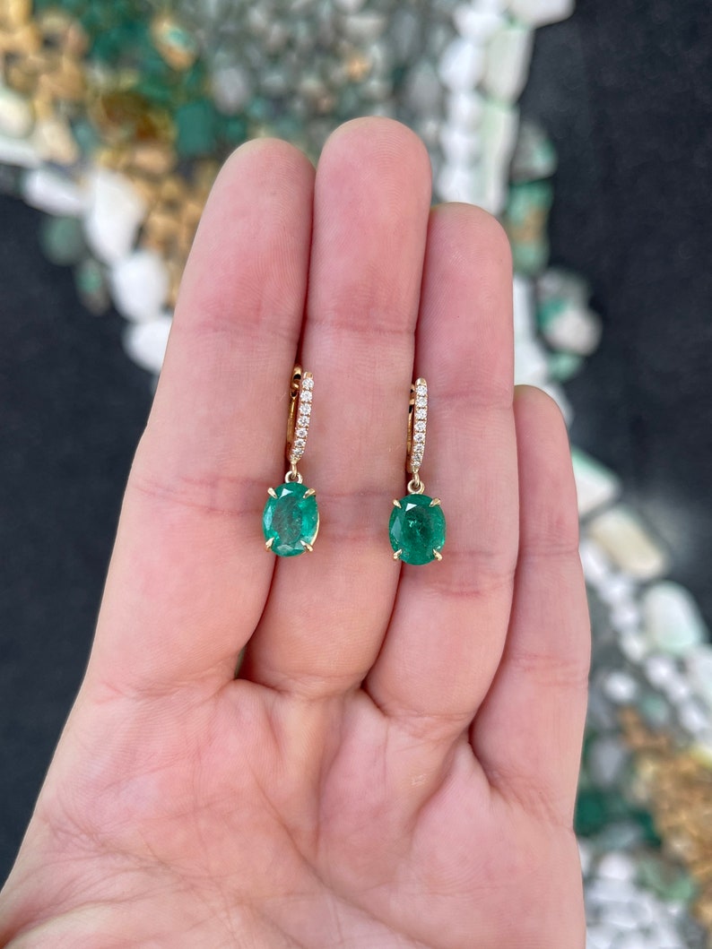 14K gold huggie dangle earrings featuring dark green oval cut emerald and diamond accents.