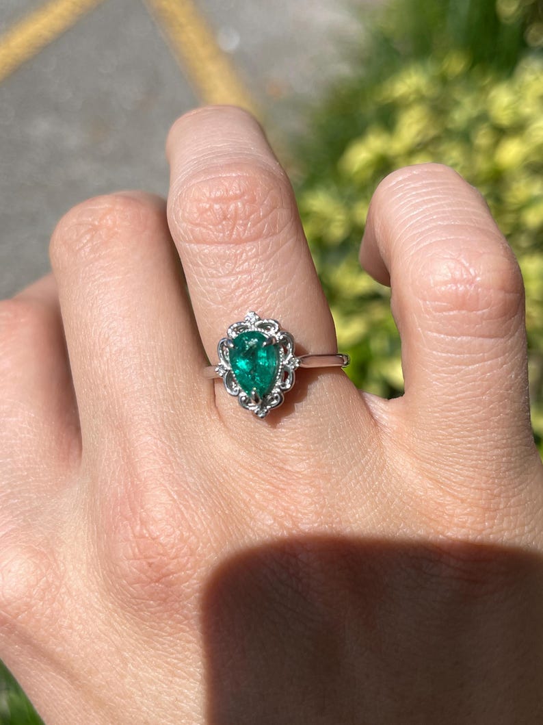 Platinum engagement ring with pear-shaped emerald and diamond accents, featuring intricate filigree crown scroll design.