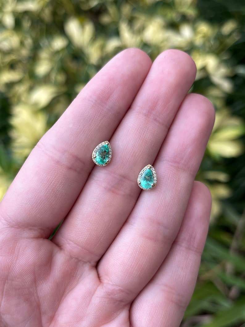 Colombian Emerald and diamond halo pear cut earrings in 14K gold featuring 1.07 carats of natural emeralds and sparkling diamonds.