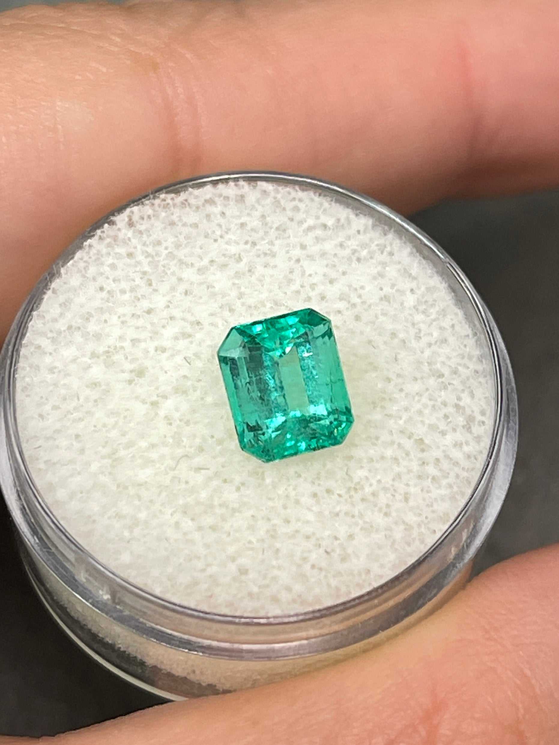 Bright bluish green 1.69 carat Colombian emerald loose gemstone in an emerald cut, displayed next to a gold ring setting.